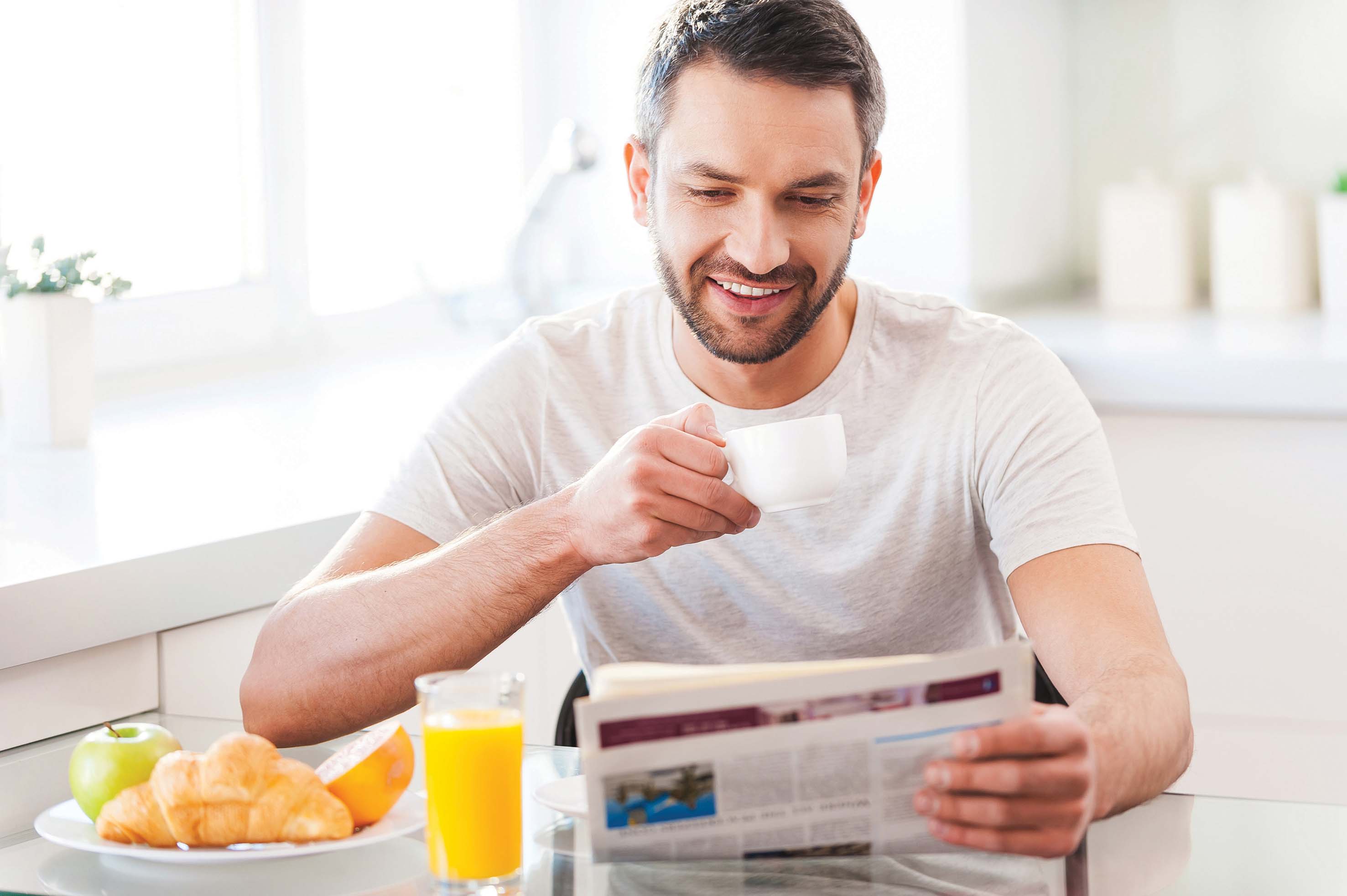 Starting day from good news. Handsome young man reading newspaper and smiling while drinking coffee and having breakfast in the kitchen